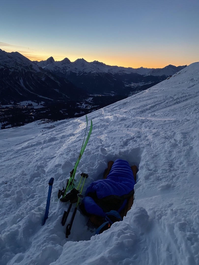 Berggipfelkette mit Sonnenaufgangsstimmung im Winter , im Vordergrund ein Loch im Schnee mit blauem Schlafsack und Ski